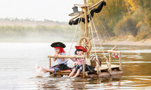 Children In Pirate Costumes Play On A Wooden Raft At Sunset. Two Girls Pretend To Be The Captains Of A Ship With Black Sails And A Flag. Funny Kids Dreaming About Adventure And Travel.