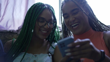 Wall Mural - Two young black women laughing and smiling holding phone. Young women looking at smartphone device