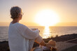 © luciano - Back view of relaxed and romantic senior woman or pensioner sitting admiring the sea at sunset light looking at the horizon - old female  outdoors enjoying vacations