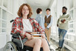 © pressmaster - Smiling teenage girl with physical disability in casualwear sitting in wheelchair in college corridor against group of classmates
