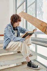 Wall Mural - Serious youthful girl in casualwear sitting on staircase in college corridor and reading book while preparing for lesson or exam at break