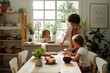 © pressmaster - Young woman making sandwiches for youthful daughter and herself for breakfast while standing by kitchen table next to girl eating