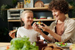 © pressmaster - Young laughing brunette woman looking at vegetable sandwich in her hands while sitting by served table next to her youthful daughter