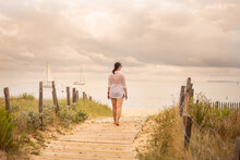 A Young Woman Walks On The Wooden Pontoon That Leads To The Beach. In The Background You Can See Two Boats With White Sails, Sailing In The Calm Water Of The Ocean. The Fence Forms A Path To The Beach