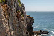 Brownstone Cliffs On The Portuguese Atlantic Coast On A Summer Day With Some Clouds And People Climbing With Ropes And Helmets