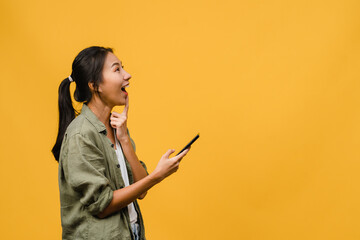 Wall Mural - Young Asia lady using phone with positive expression, smiles broadly, dressed in casual clothing feeling happiness and stand isolated on yellow background. Happy adorable glad woman rejoices success.