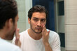 © Vitaliy - Handsome man looking at mirror and applying moisturizing cream on cheeks in bathroom. Groomed young guy doing skincare morning routine after taking a shower