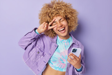 Wall Mural - positive carefree young woman with curly hair makes peace gesture over eye smiles happily holds mobi