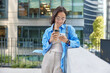 © wayhome.studio  - Pretty Asian woman watches video in social networks on smartphone drinks takeaway coffee sends text messages wears spectacles and casual clothes poses against urban building. Modern lifestyle