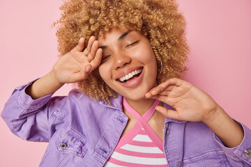 Sticker - Happy dreamy woman with curly blonde hair smiles broadly keeps eyes closed touches face gently recalls pleasant memories enjoys life wears fashionable purple jacket isolated over pink background