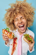 Photo of curly haired woman with curly hair holds delicious hot dog and burger enjoys eating fast food dressed in white formal shirt and tie smeared with ketchup isolated over blue background