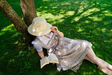 a woman wearing a hat and a beautiful traditional Bavarian or also Austrian dirndl dress called Tracht resting with a good book on the green meadow on a sunny summer day (Munich, Bavaria, Germany)