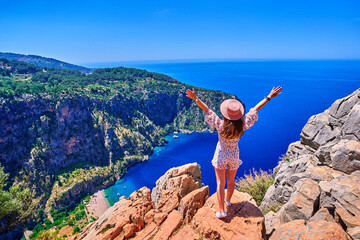 Wall Mural - Free carefree joyful girl traveler with open arms stands on hill rock over sea bay in Turkey, butterfly valley. Enjoy travel concept