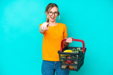 Blonde English young girl holding a shopping basket full of food isolated on blue background surprised and pointing front