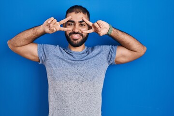 Sticker - Middle east man with beard standing over blue background doing peace symbol with fingers over face, smiling cheerful showing victory