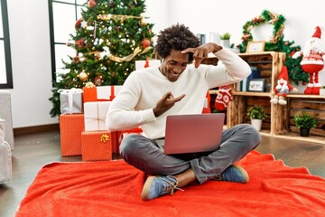 Sticker - Young african american man using laptop sitting by christmas tree smiling making frame with hands and fingers with happy face. creativity and photography concept.