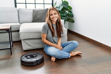 Canvas Print - Young caucasian woman sitting at home by vacuum robot looking positive and happy standing and smiling with a confident smile showing teeth