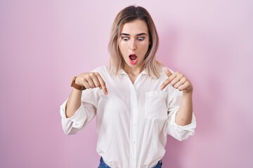 Canvas Print - Young beautiful woman standing over pink background pointing down with fingers showing advertisement, surprised face and open mouth