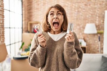 Sticker - Young beautiful woman holding led lightbulb and incandescent bulb angry and mad screaming frustrated and furious, shouting with anger looking up.