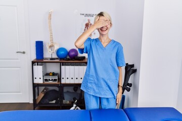 Poster - Young physiotherapist woman working at pain recovery clinic smiling and laughing with hand on face covering eyes for surprise. blind concept.