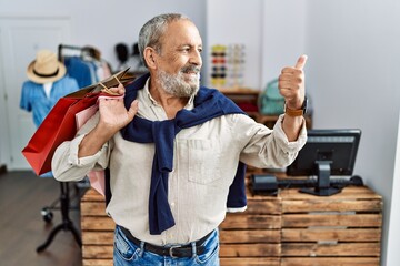 Canvas Print - Handsome senior man holding shopping bags at boutique shop smiling with happy face looking and pointing to the side with thumb up.