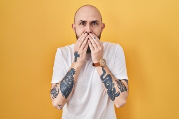 Poster - Young hispanic man with tattoos standing over yellow background laughing and embarrassed giggle covering mouth with hands, gossip and scandal concept