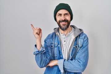 Canvas Print - Young hispanic man with tattoos wearing wool cap with a big smile on face, pointing with hand and finger to the side looking at the camera.