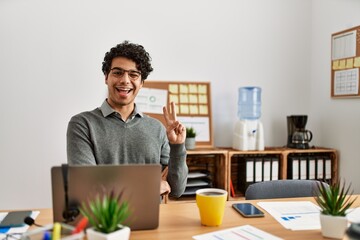 Canvas Print - Young hispanic man wearing business style sitting on desk at office smiling with happy face winking at the camera doing victory sign. number two.