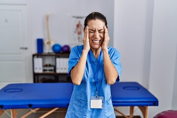 Wall Mural - Young hispanic woman wearing physiotherapist uniform standing at clinic with hand on head for pain in head because stress. suffering migraine.