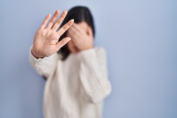 Poster - Young brunette woman standing over blue background covering eyes with hands and doing stop gesture with sad and fear expression. embarrassed and negative concept.