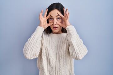 Sticker - Young brunette woman standing over blue background trying to open eyes with fingers, sleepy and tired for morning fatigue