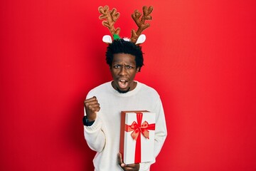 Canvas Print - Young african american man wearing deer christmas hat holding gift angry and mad raising fist frustrated and furious while shouting with anger. rage and aggressive concept.