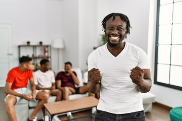 Poster - Young african man with friends at the living room very happy and excited doing winner gesture with arms raised, smiling and screaming for success. celebration concept.