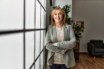 Wall Mural - Middle age businesswoman standing with arms crossed gesture leaning on the window at the office.