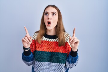 Wall Mural - Young hispanic girl standing over blue background amazed and surprised looking up and pointing with fingers and raised arms.