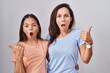 Young mother and daughter standing over white background surprised pointing with hand finger to the side, open mouth amazed expression.