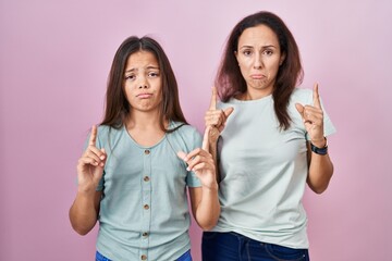 Wall Mural - Young mother and daughter standing over pink background pointing up looking sad and upset, indicating direction with fingers, unhappy and depressed.