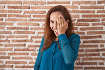 Canvas Print - Brunette woman standing over bricks wall covering one eye with hand, confident smile on face and surprise emotion.