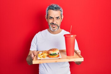 Sticker - Handsome middle age man with grey hair eating a tasty classic burger with fries and soda clueless and confused expression. doubt concept.