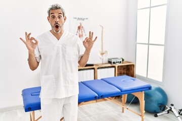 Poster - Middle age hispanic therapist man working at pain recovery clinic looking surprised and shocked doing ok approval symbol with fingers. crazy expression