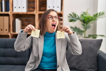 Wall Mural - Middle age hispanic woman working on depression holding sad to happy emotion paper angry and mad screaming frustrated and furious, shouting with anger. rage and aggressive concept.