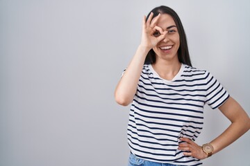 Sticker - Young brunette woman wearing striped t shirt doing ok gesture with hand smiling, eye looking through fingers with happy face.