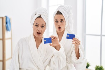 Sticker - Middle age woman and daughter wearing bathrobe holding credit card scared and amazed with open mouth for surprise, disbelief face