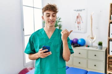 Wall Mural - Young caucasian physio man using smartphone at medical clinic pointing thumb up to the side smiling happy with open mouth