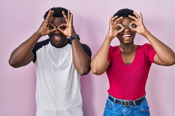 Poster - Young african american couple standing over pink background doing ok gesture like binoculars sticking tongue out, eyes looking through fingers. crazy expression.