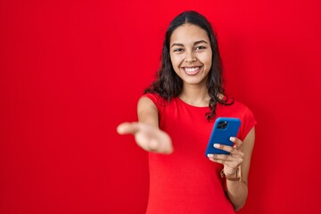 Poster - Young brazilian woman using smartphone over red background smiling cheerful offering palm hand giving assistance and acceptance.