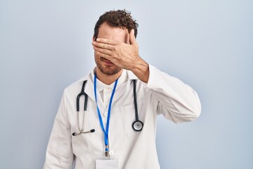 Sticker - Young hispanic man wearing doctor uniform and stethoscope smiling and laughing with hand on face covering eyes for surprise. blind concept.