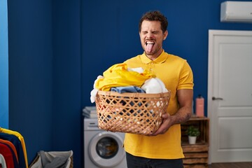 Wall Mural - Young hispanic man holding laundry basket sticking tongue out happy with funny expression.