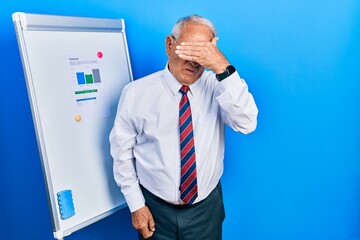 Canvas Print - Senior man with grey hair standing by business blackboard covering eyes with hand, looking serious and sad. sightless, hiding and rejection concept