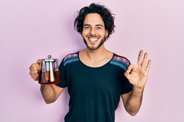 Wall Mural - Handsome hispanic man holding traditional tea pot doing ok sign with fingers, smiling friendly gesturing excellent symbol